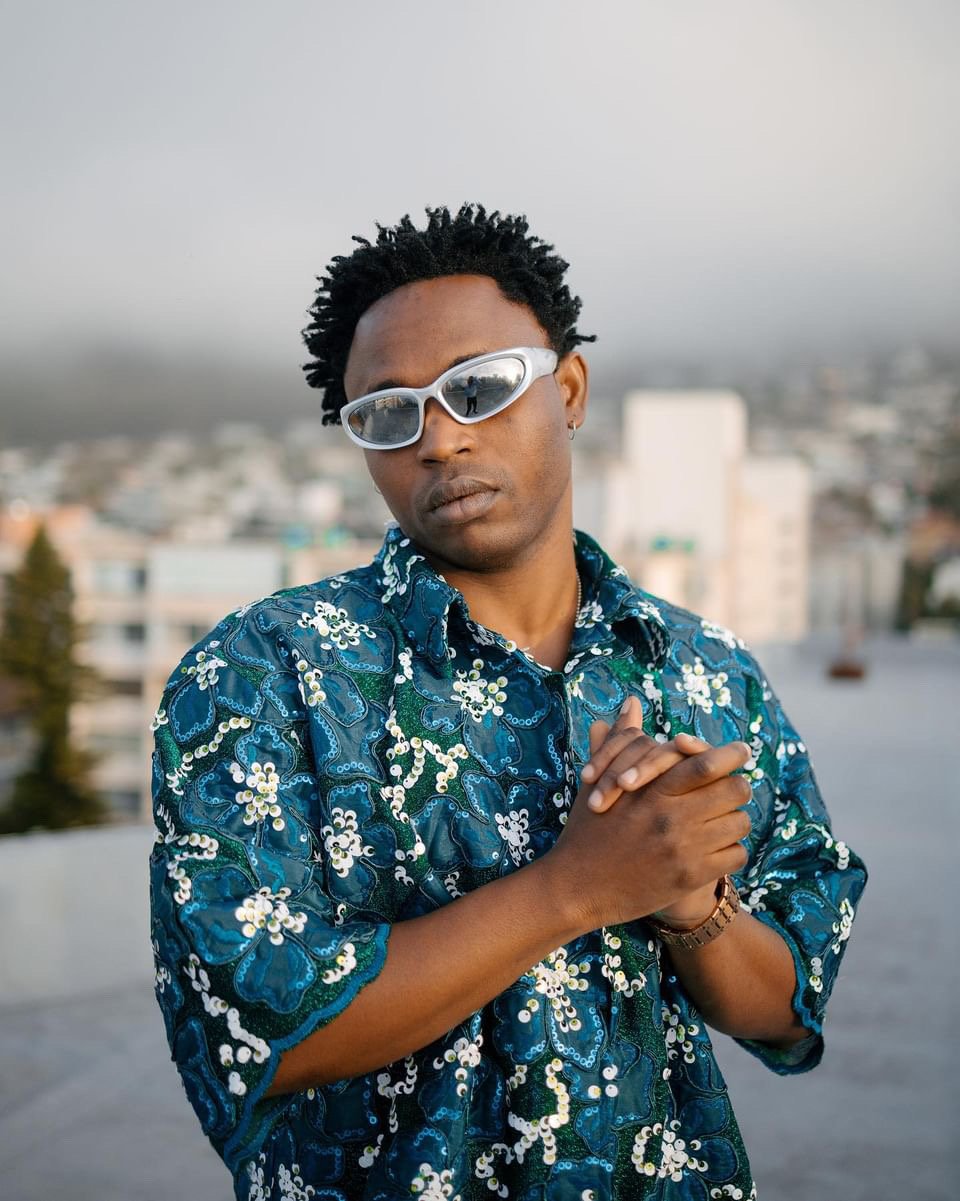Demmie Vee on the rooftop, teal floral shirt and white sunglasses, city behind him.