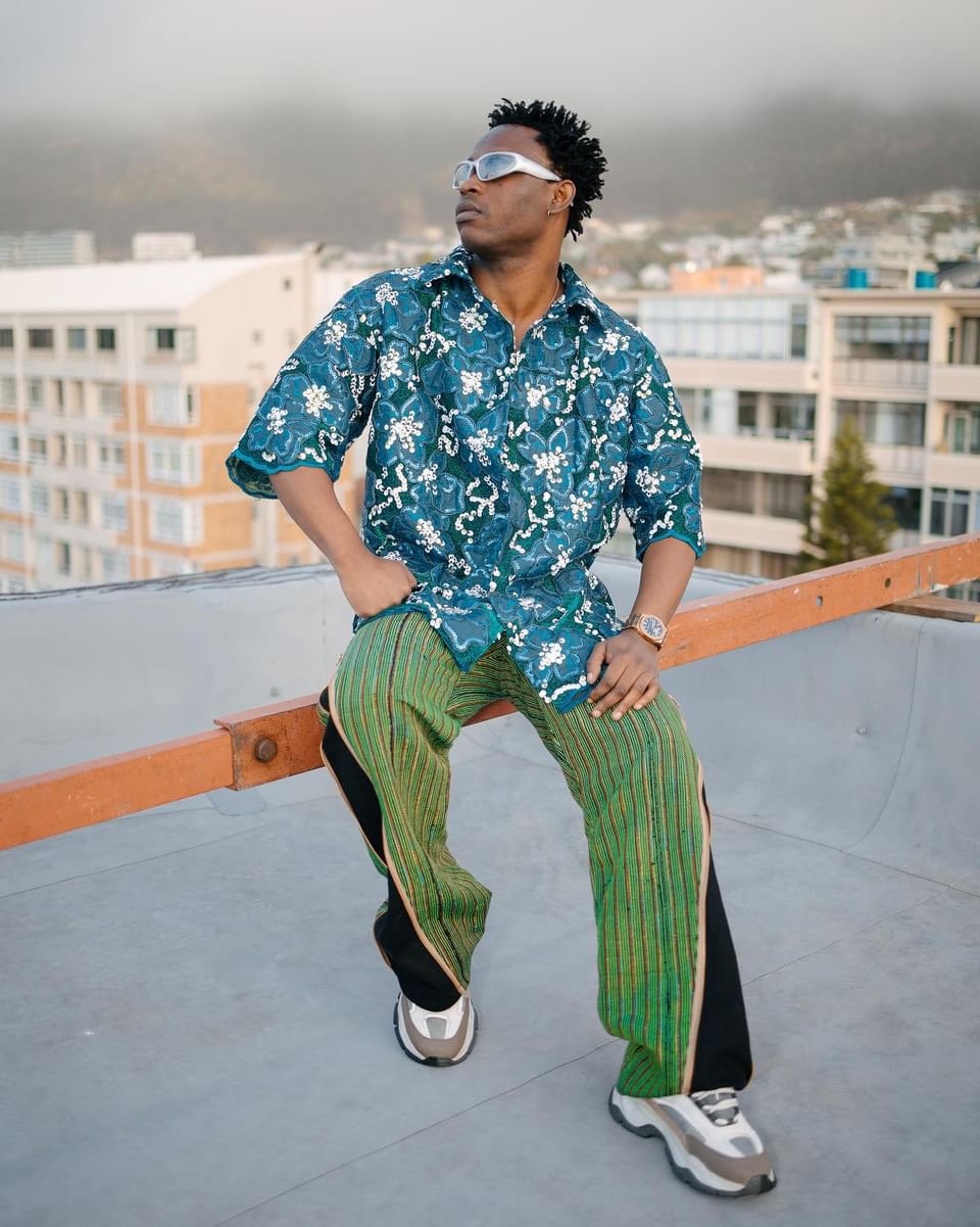 Demmie Vee in green trousers on a Lagos rooftop, skyline in the background.