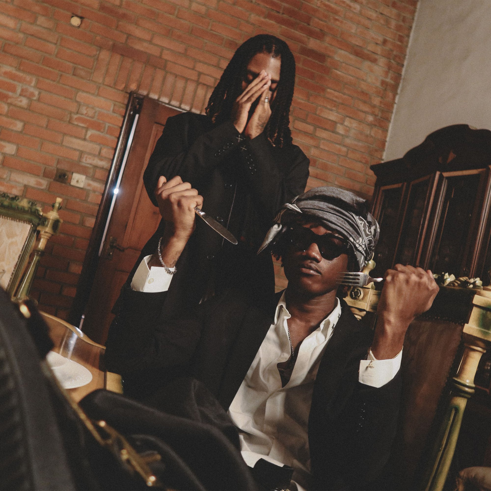 A still from the Big Drop video: two men at a dining table against a brick wall, one standing with a knife over his face, one seated in a grey headwrap and sunglasses.